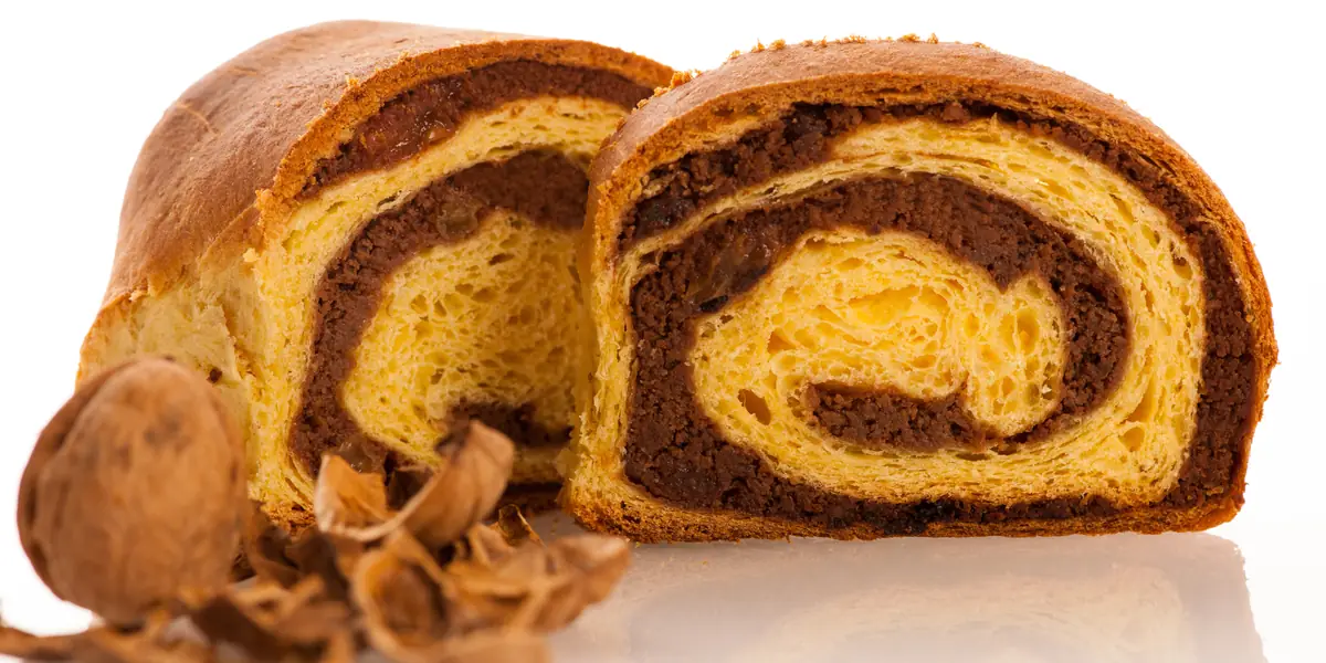 Cozonac cu nucă tăiat în două, cu spirala de umplutură de nucă și cacao la vedere, lângă o nucă și coji