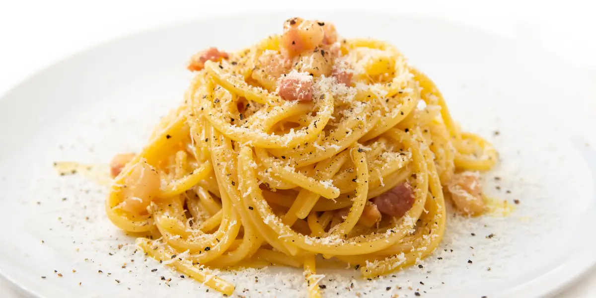 Spaghete carbonara pe o farfurie albă, cu bucăți de guanciale, pecorino ras și piper negru