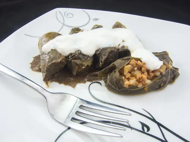 Sarmale în foi de viță pe o farfurie albă, cu iaurt deasupra, una tăiată cu umplutura de carne și orez la vedere