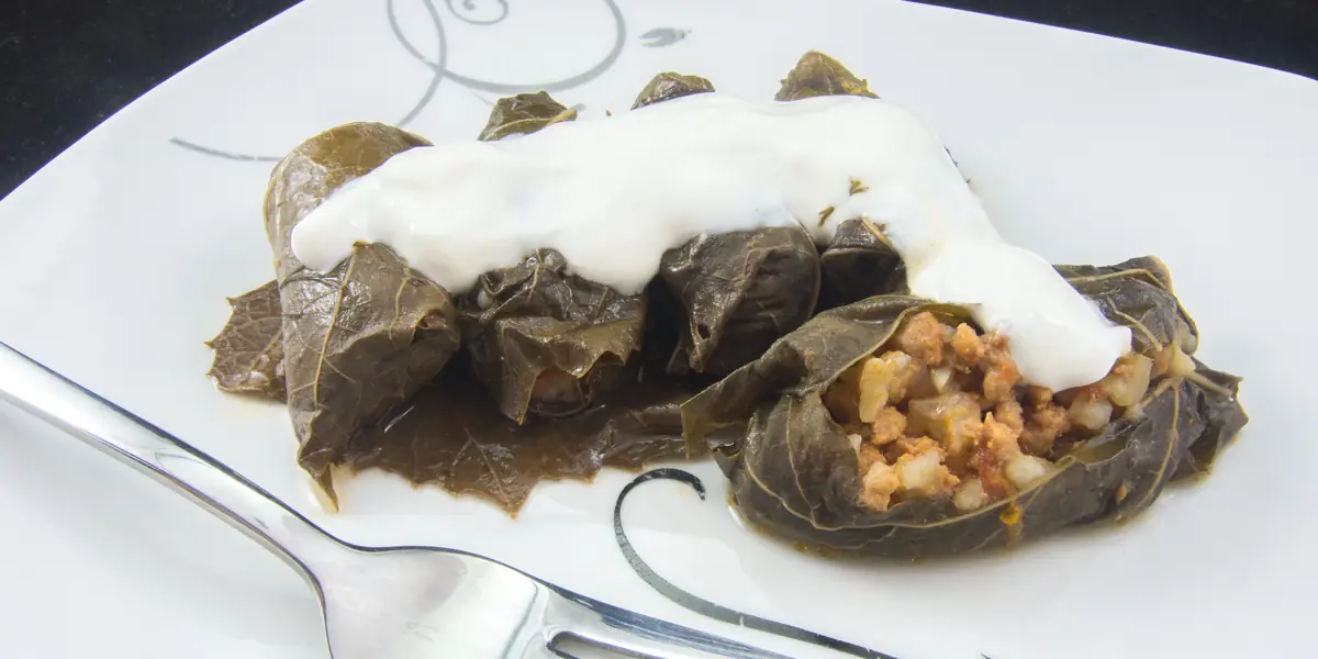 Sarmale în foi de viță pe o farfurie albă, cu iaurt deasupra, una tăiată cu umplutura de carne și orez la vedere