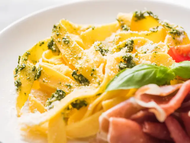 Tagliatelle cu pesto verde și parmezan ras, cu felii de prosciutto crudo, roșii și o frunză de busuioc, pe o farfurie albă