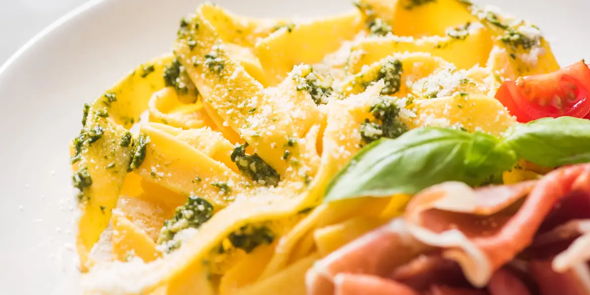 Tagliatelle cu pesto verde și parmezan ras, cu felii de prosciutto crudo, roșii și o frunză de busuioc, pe o farfurie albă