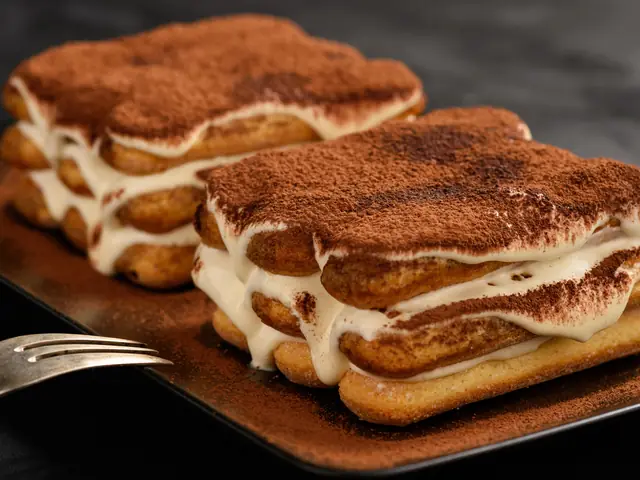 Două porții de tiramisu pe un platou închis la culoare, cu straturi de pișcoturi, cremă de mascarpone și cacao pudrată deasupra