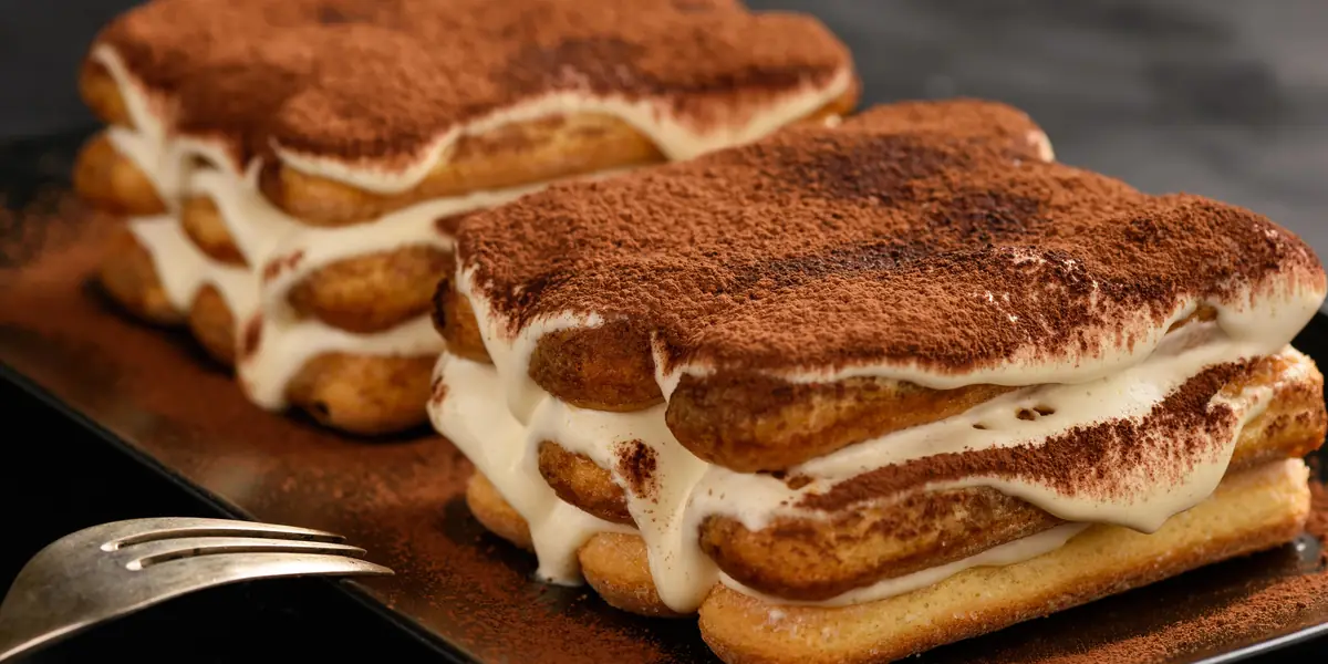 Două porții de tiramisu pe un platou închis la culoare, cu straturi de pișcoturi, cremă de mascarpone și cacao pudrată deasupra